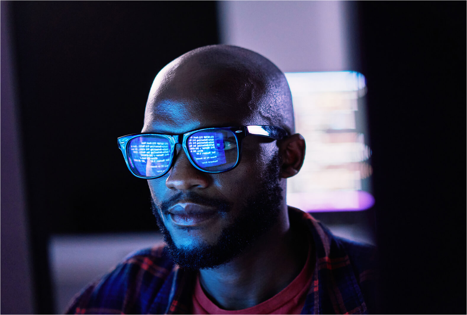 Developer wearing glasses with code reflected in the lenses while working at a computer screen in a dark room.