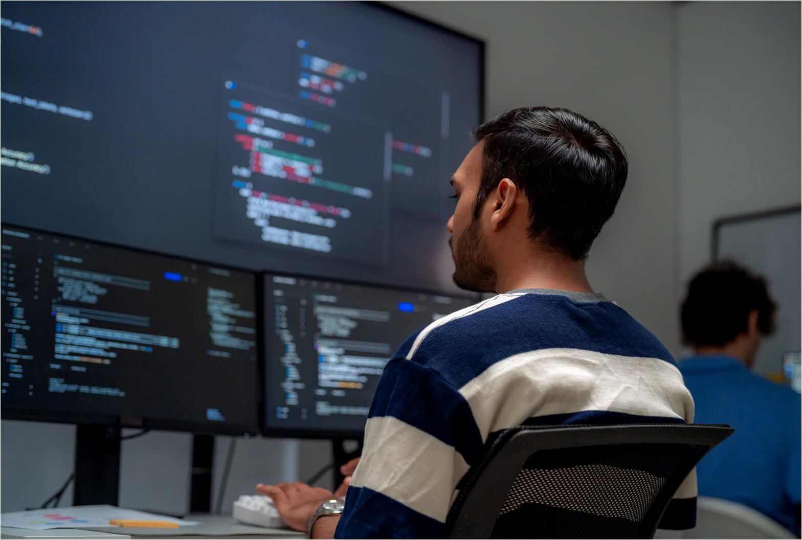 Developer in a striped sweater reviewing code on multiple monitors in an office environment.