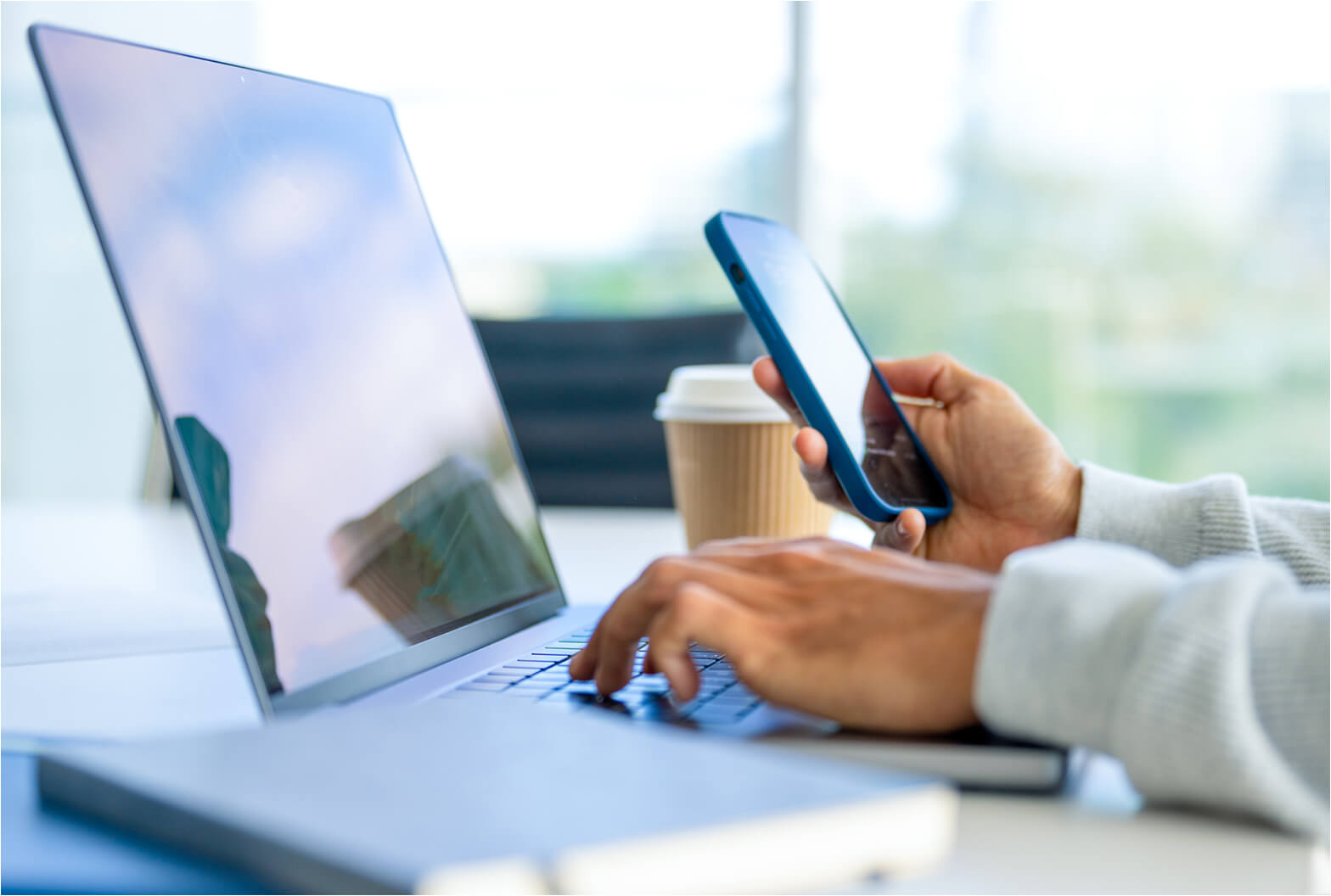 Person typing on a laptop while holding a blue smartphone, with a coffee cup on the desk near a bright window.