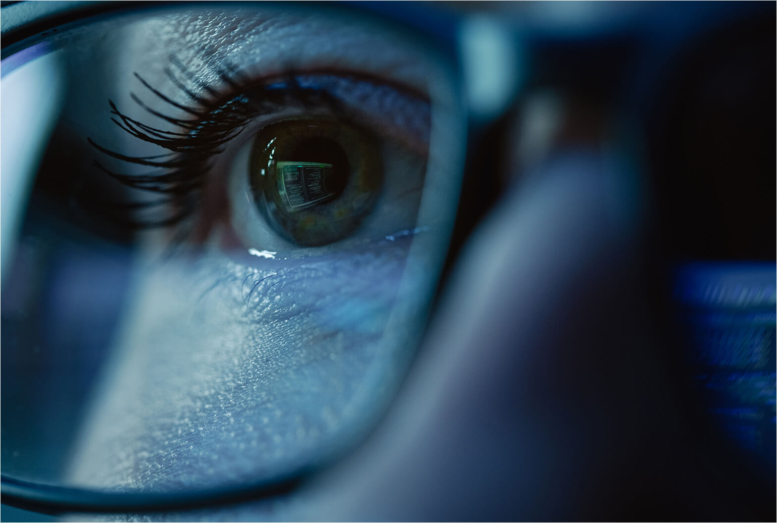 Close-up of a human eye behind glasses with a digital screen reflected in the iris, bathed in blue light.