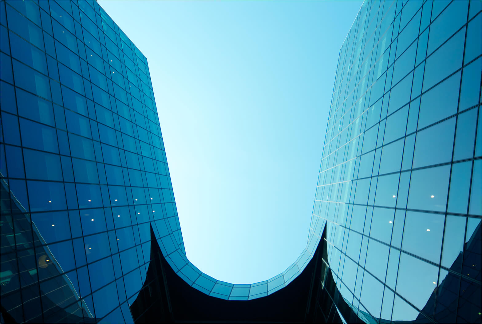 Upward view of a curved glass skyscraper with blue-tinted facades framing a clear sky between two sweeping wings.