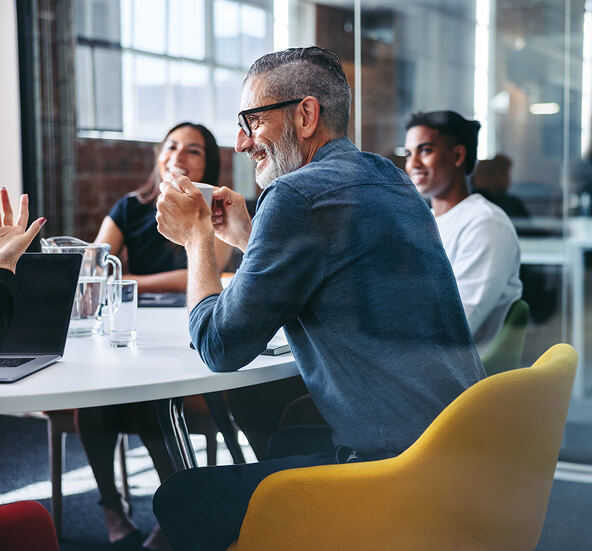 Smiling bearded man with glasses engages in a lively discussion with colleagues around a modern conference table.