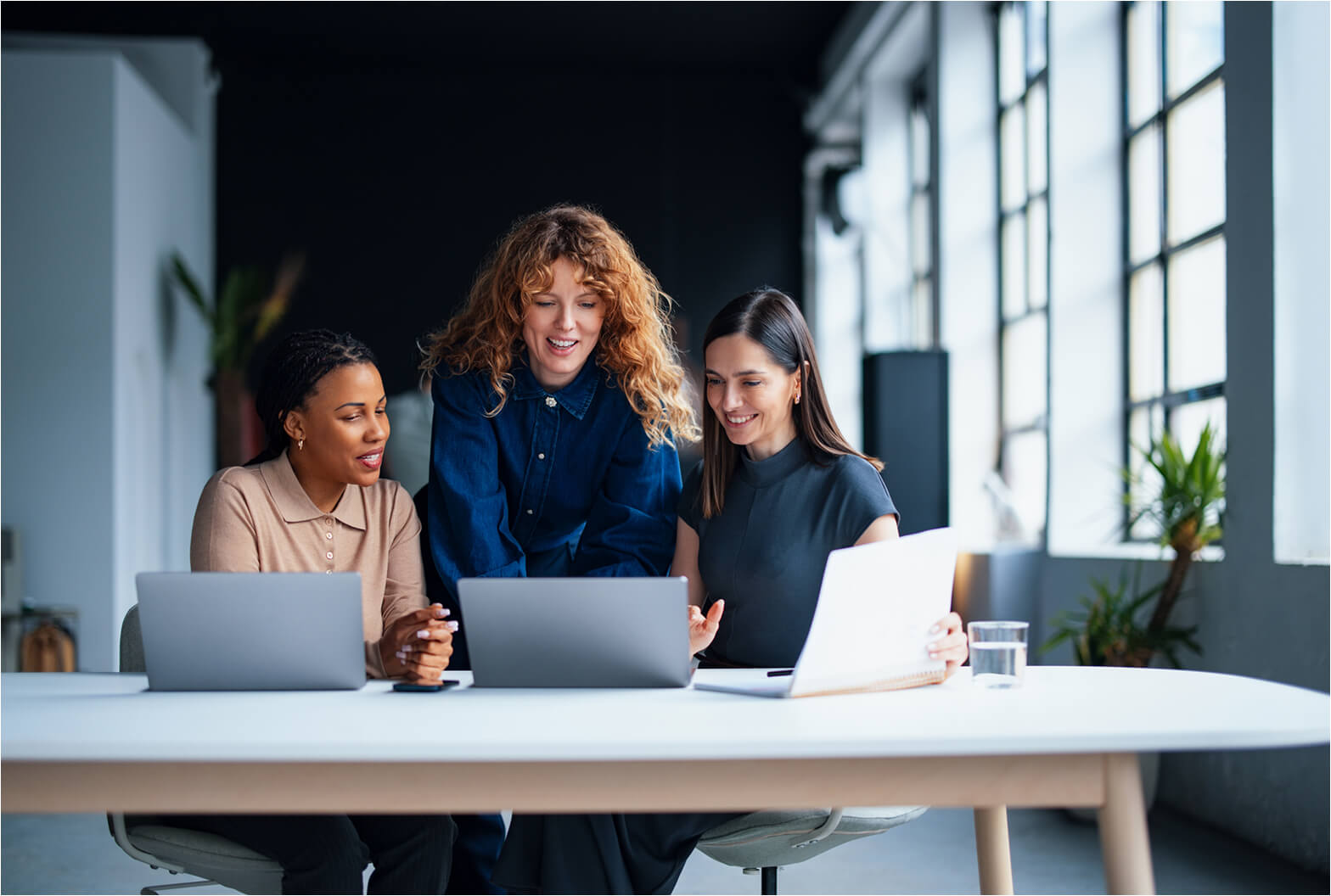 Three women collaborating at a white desk with laptops in a modern, sunlit office space with large windows.