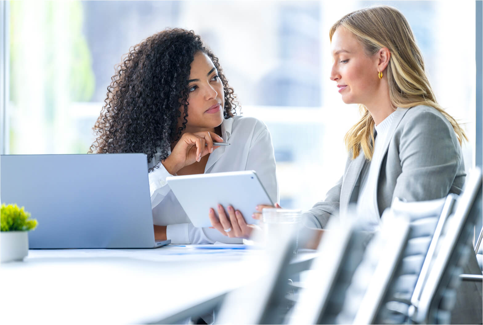 Two professional women collaborating at a bright office table with a laptop and tablet, engaged in discussion.