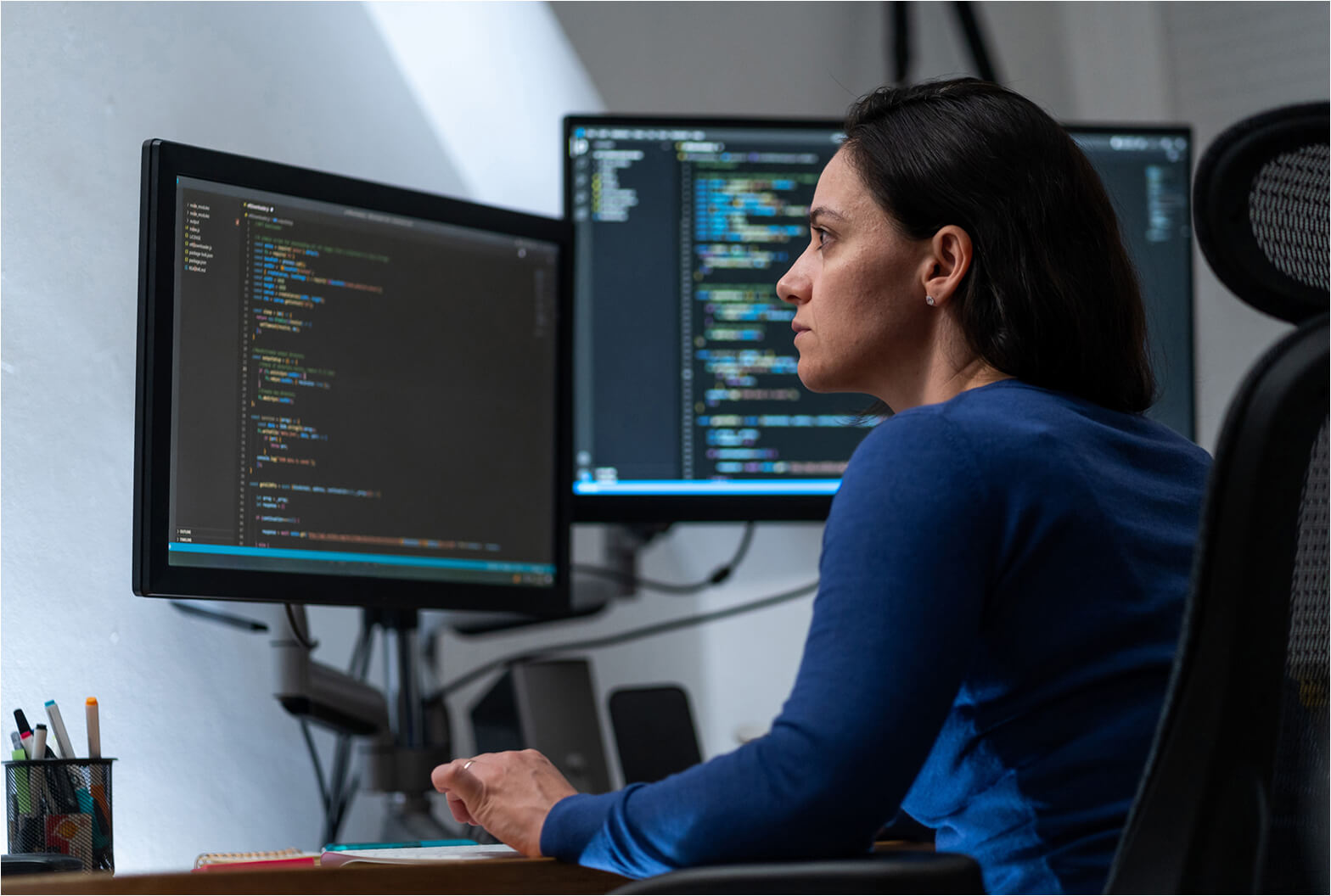 A woman in a blue sweater reviews lines of code on dual monitors at her desk in a dimly lit office.