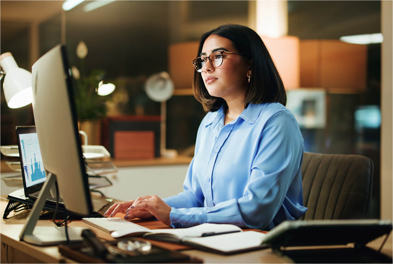 Professional woman wearing glasses reviews data charts on a desktop monitor at her office desk.
