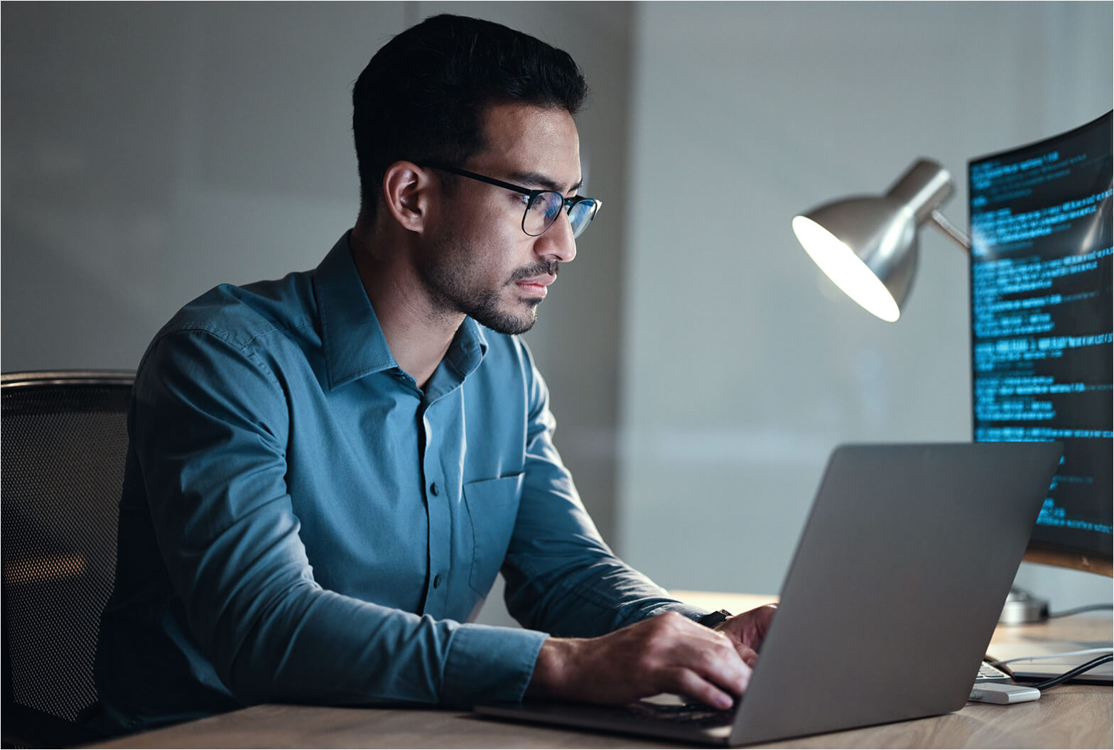 Focused man wearing glasses works on a laptop at a desk with code displayed on a monitor behind him.