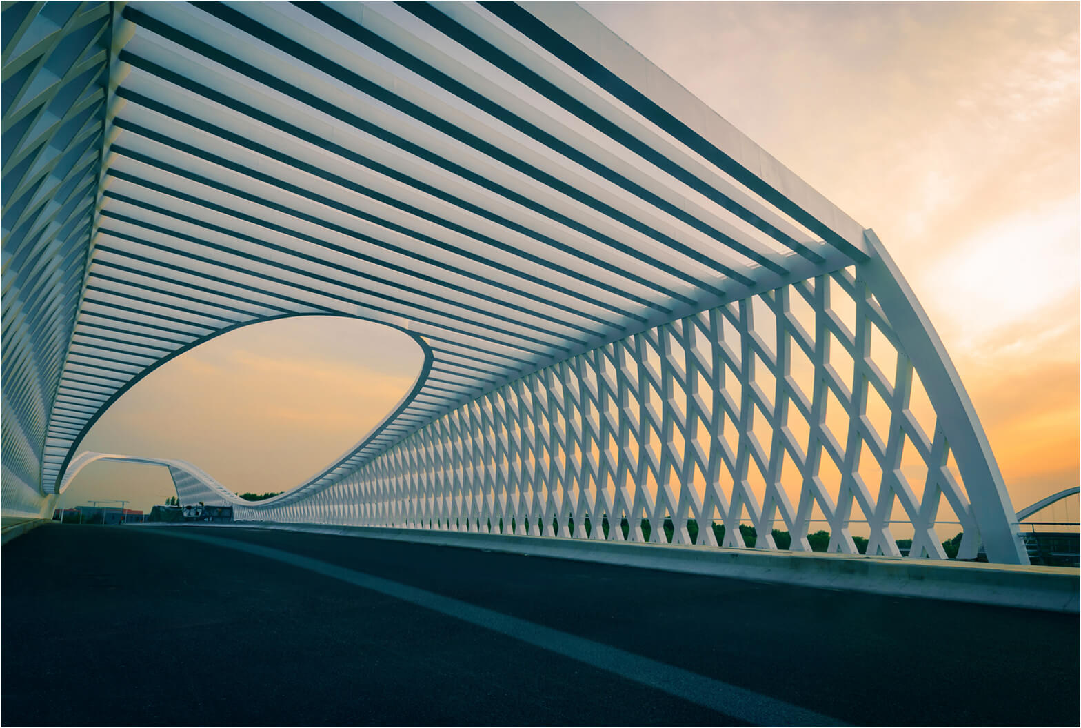 Modern white lattice bridge with sweeping curved architecture stretching into the distance against a golden sunset sky.