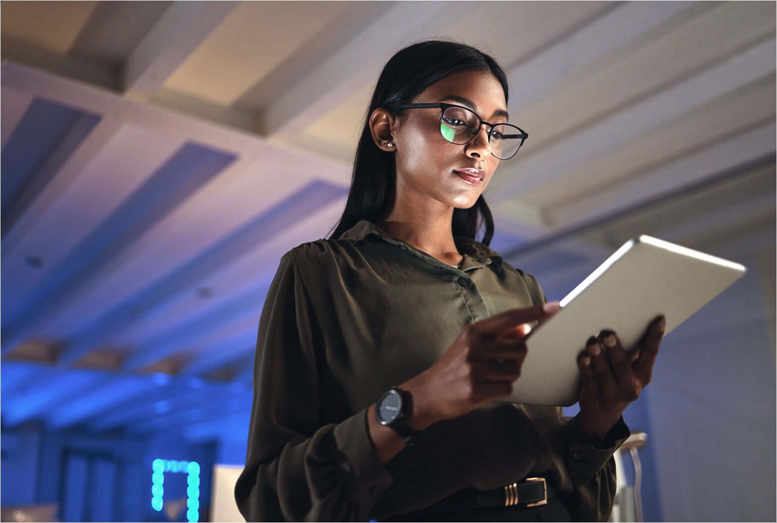 Professional woman wearing glasses reviews data on a tablet in a modern, blue-lit office environment.