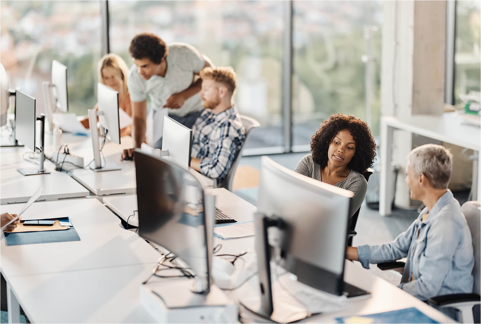 Diverse team of professionals collaborating at computer workstations in a bright, modern open-plan office with large windows.