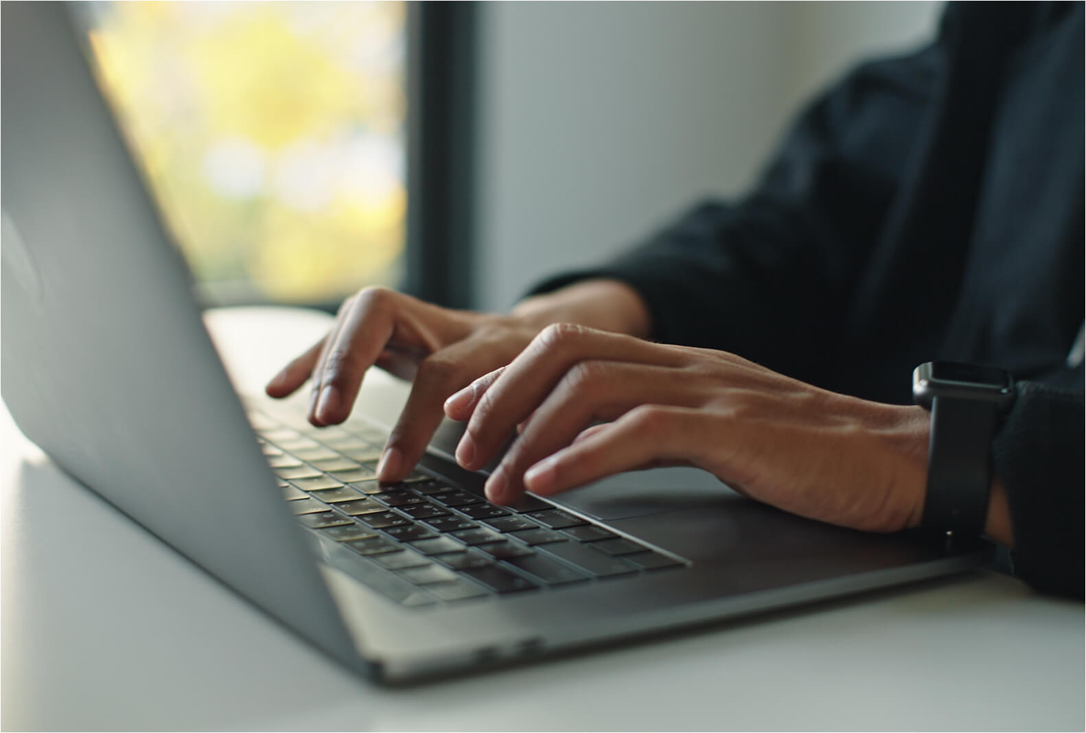 Close-up of hands typing on a silver laptop keyboard, with a smartwatch visible on the wrist.