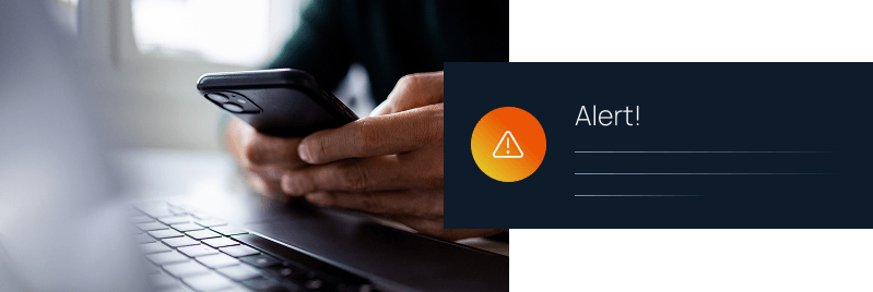Person holding a smartphone near a laptop with an orange warning icon and "Alert!" notification overlay.