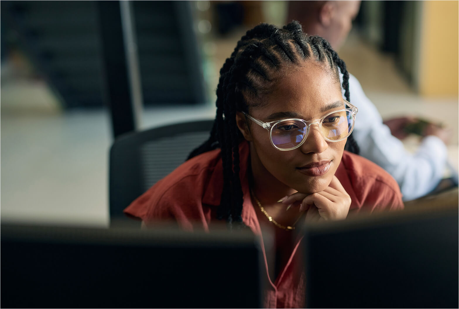 A woman with braids and glasses rests her chin on her hand while focused on a computer monitor at her desk.