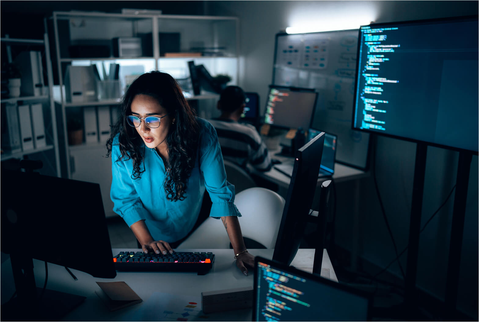 Woman wearing glasses reviews code on multiple monitors in a dimly lit office while typing on a mechanical keyboard.