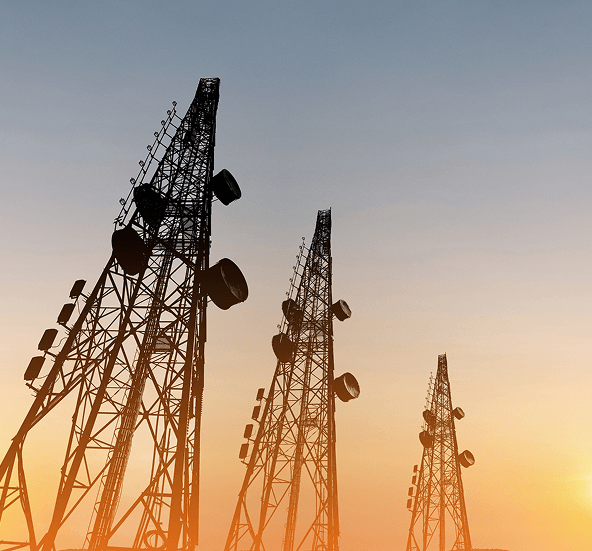 Silhouetted telecommunications towers with satellite dishes and antennas standing against a warm sunset sky.