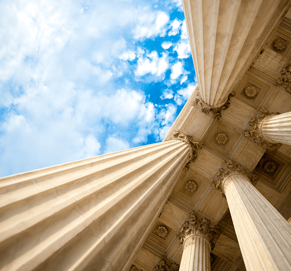 Upward view of grand marble columns and ornate ceiling of a federal government building against a bright blue sky.